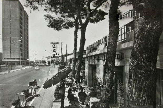 strada con un bar all'aperto, dove persone sono sedute ai tavolini sotto gli alberi.